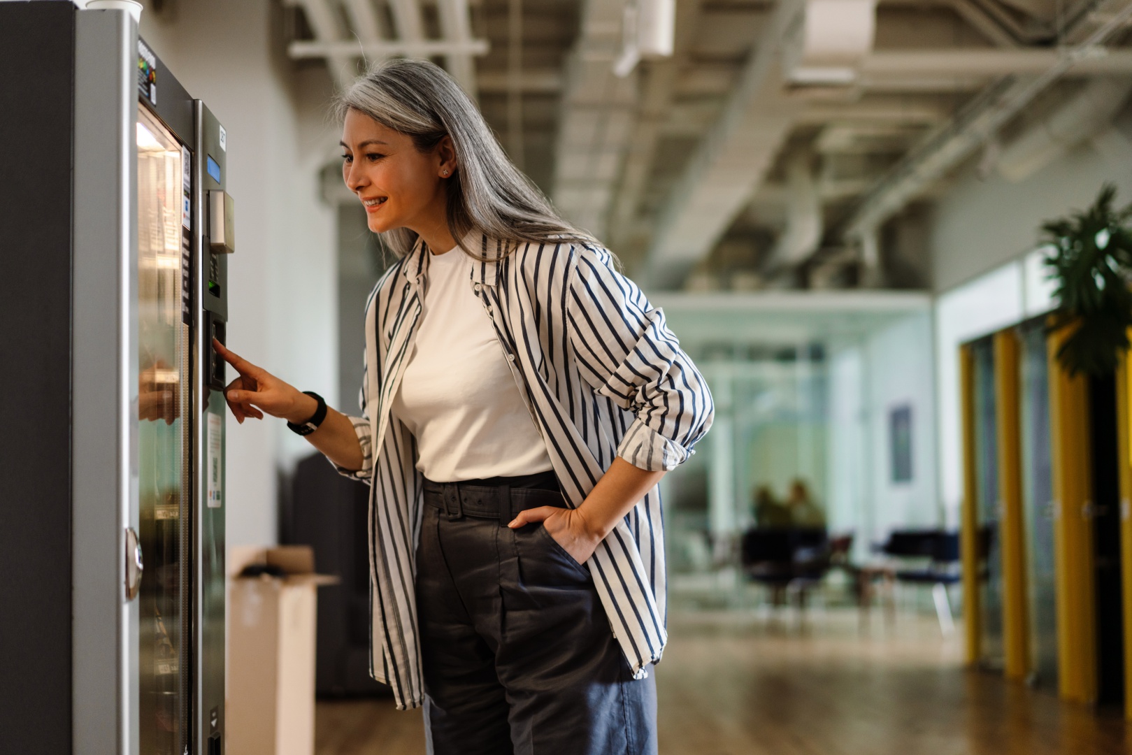 Happy white-haired mature woman using vending machine and smiling indoors